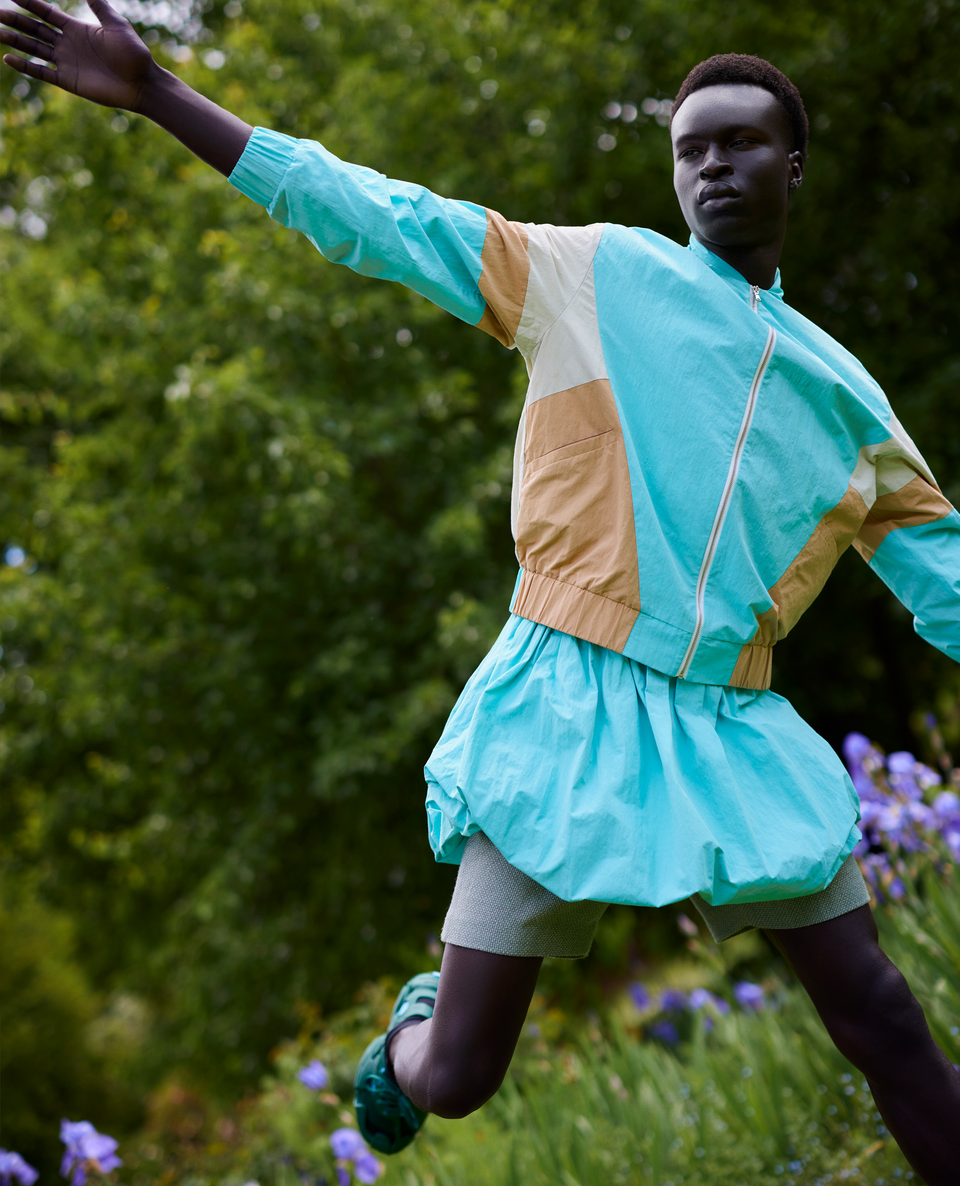 Fashion shot of Black male model outdoors in light-blue, beige, and white jacket and matching skirt, running on green grass, smiling.