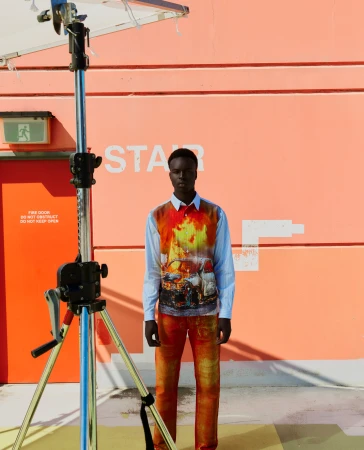 An adult Black male stands in front of a peach wall wearing an abstract printed button-down shirt and matching pants in direct sunlight.