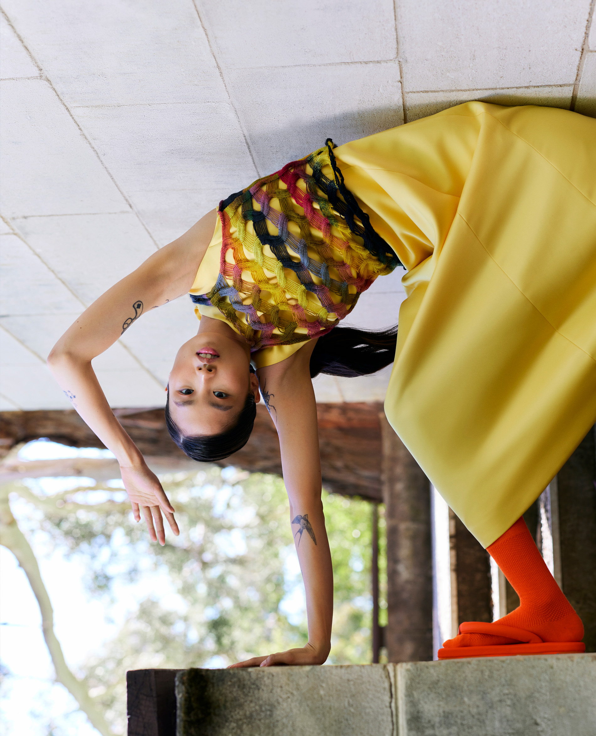 A young woman in a backbend wearing a yellow dress, orange socks and shoes stands on a concrete structure outdoors.