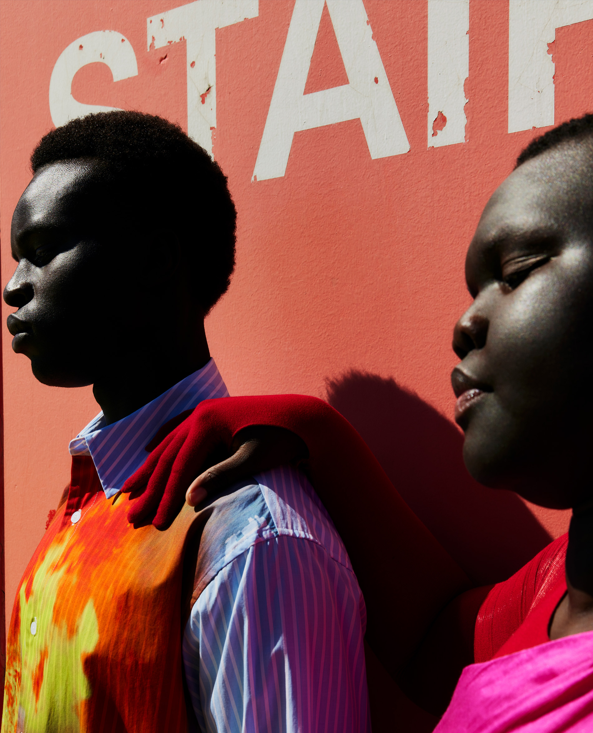 Two dark-skinned adult women pose side-by-side in front of a pink wall with white text, wearing a colorful top and pink top, in an outdoor setting.