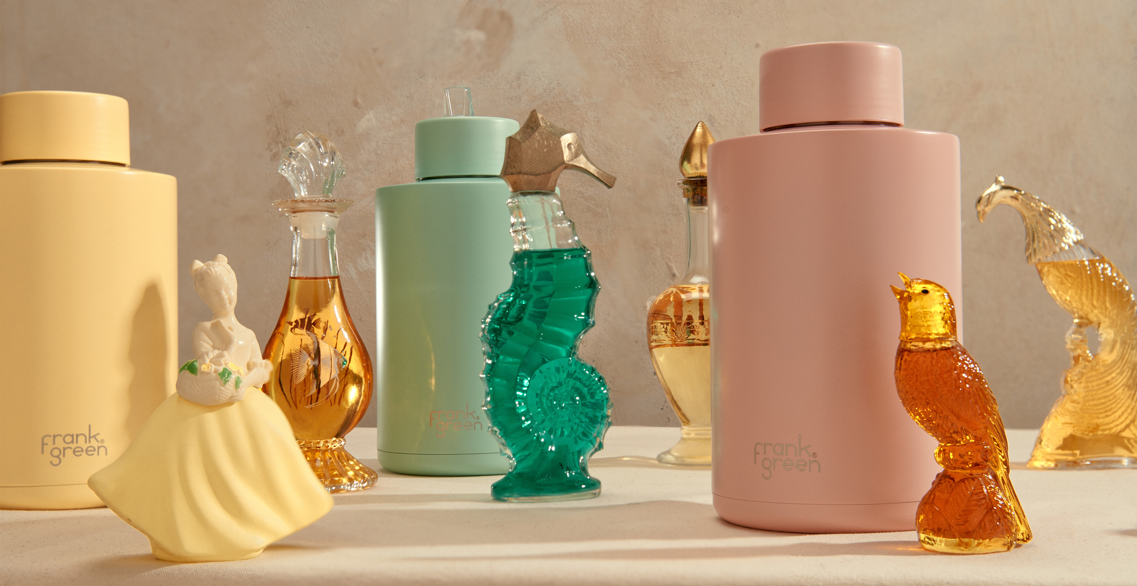 Several Frank Green water bottles in pastel colors alongside decorative glass bottles shaped like a bird, seahorse, and woman.