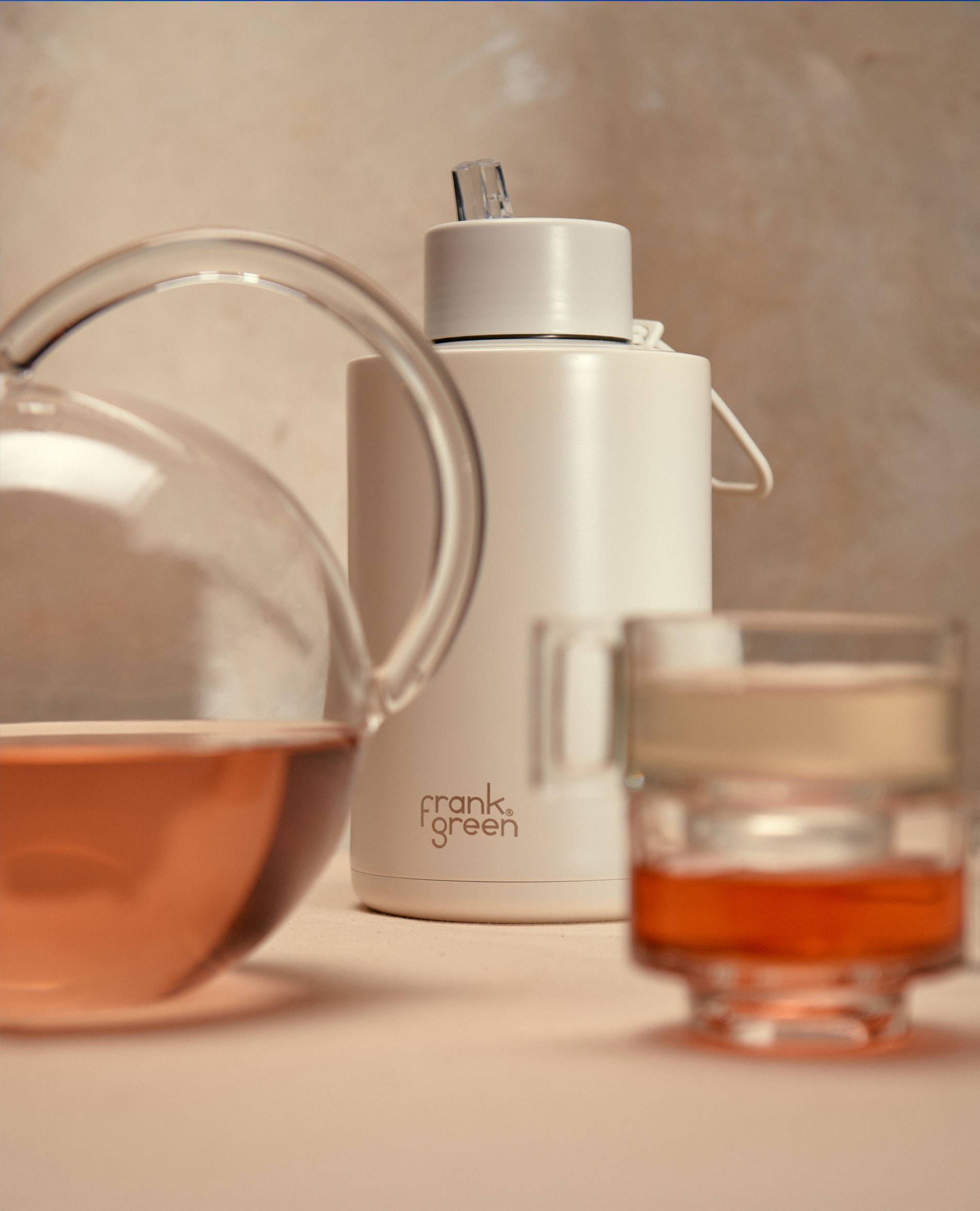 Still life featuring a glass teapot with rose-colored tea, a white water bottle with 'frank green' logo, and a glass teacup filled with tea.