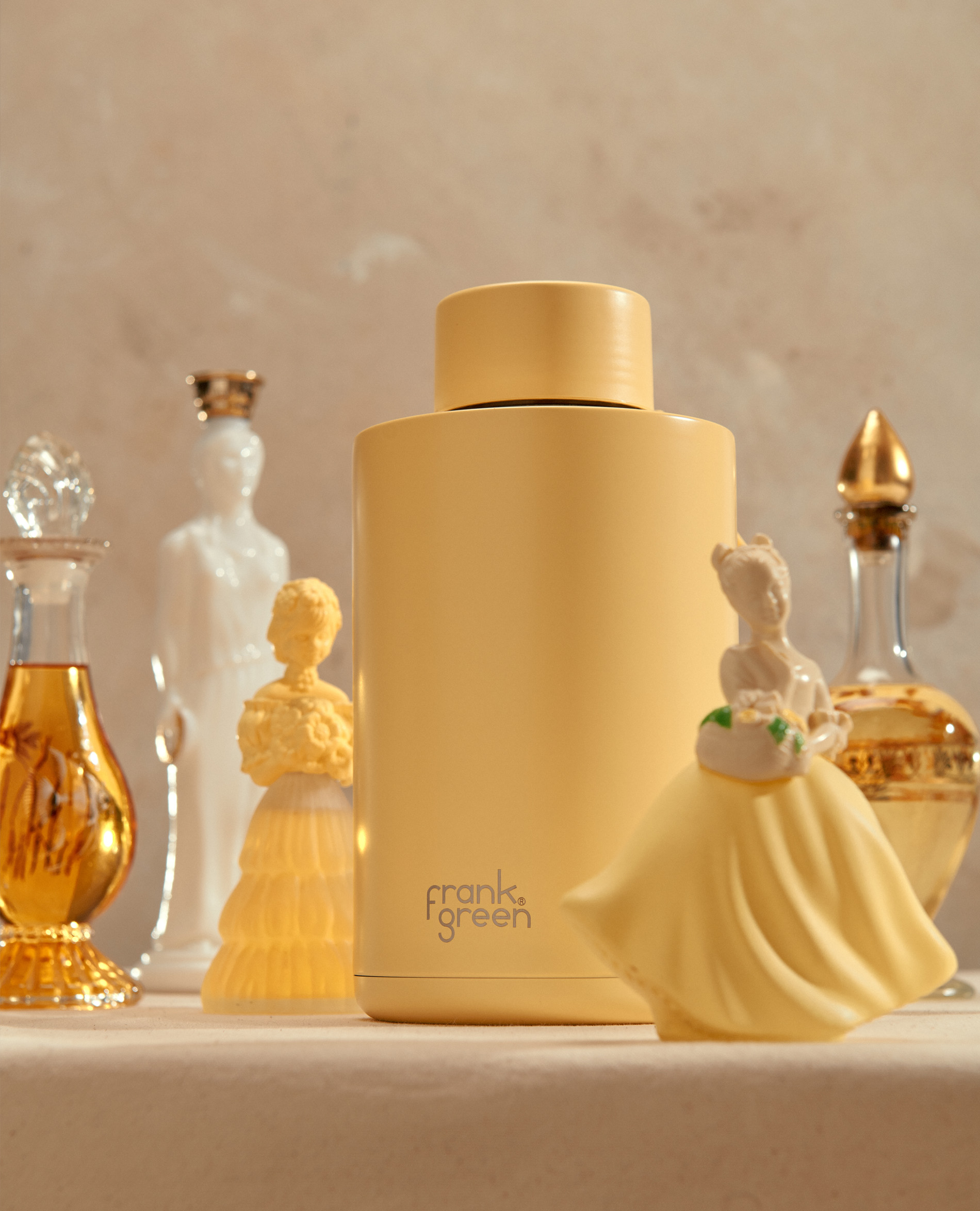Still life of a light yellow Frank Green water bottle, with vintage perfume bottles and decorative statuettes against a neutral background.