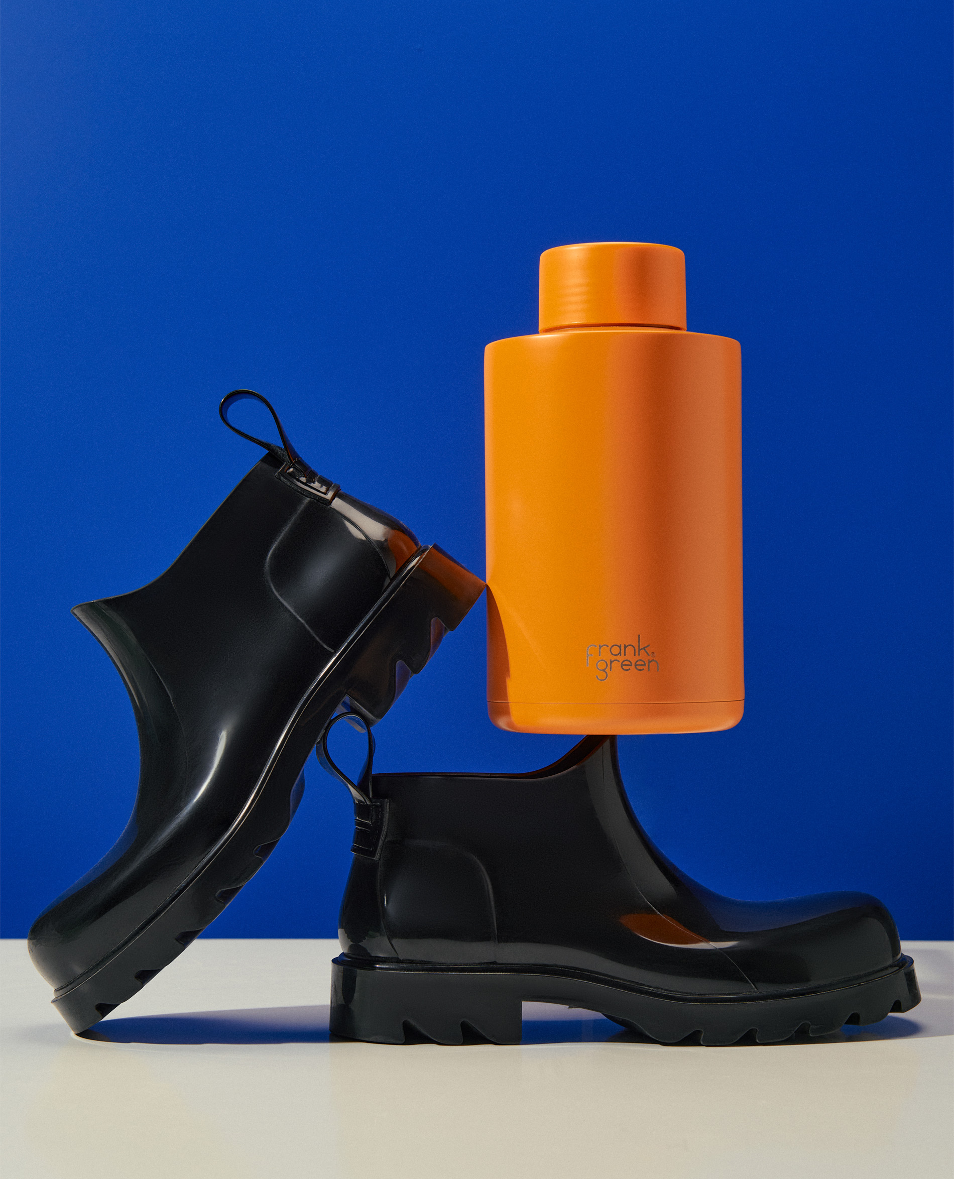 Two black rubber boots and an orange water bottle on a white surface against a blue background, creating a modern product still life.