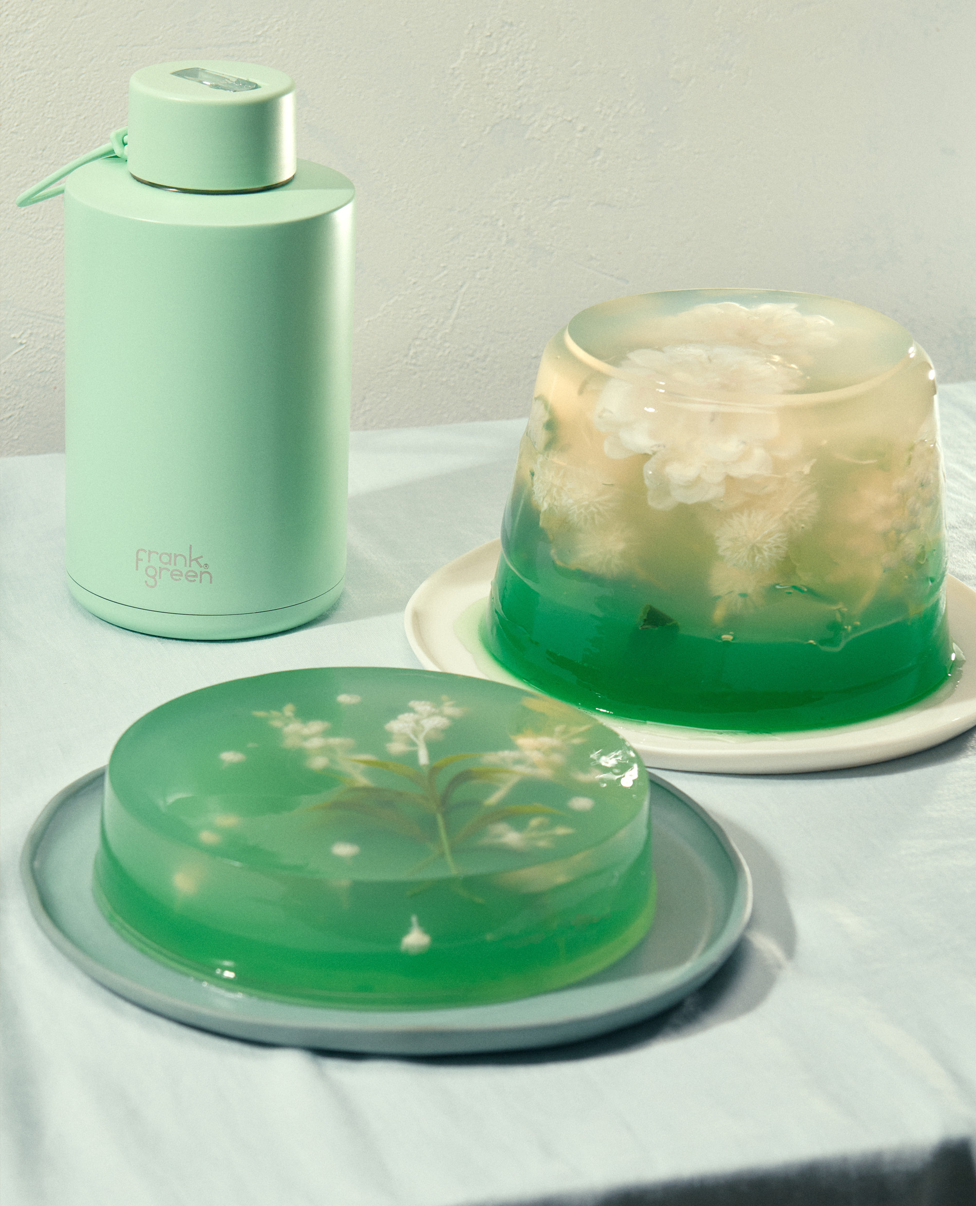 Still life of two green gelatin desserts with floral details, alongside a matching green Frank Green water bottle against a neutral background.