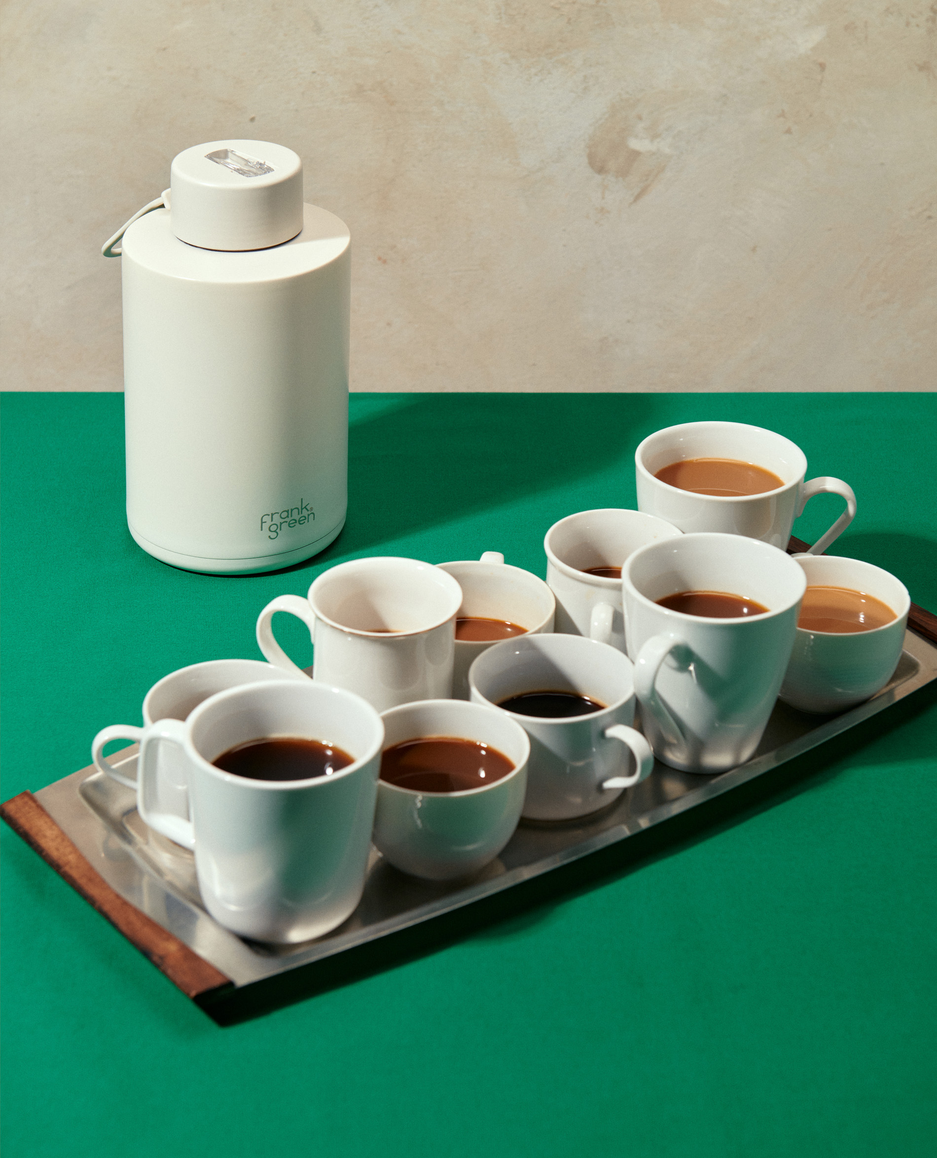 An assortment of white ceramic coffee cups filled with coffee on a silver tray alongside a white Frank Green water bottle.