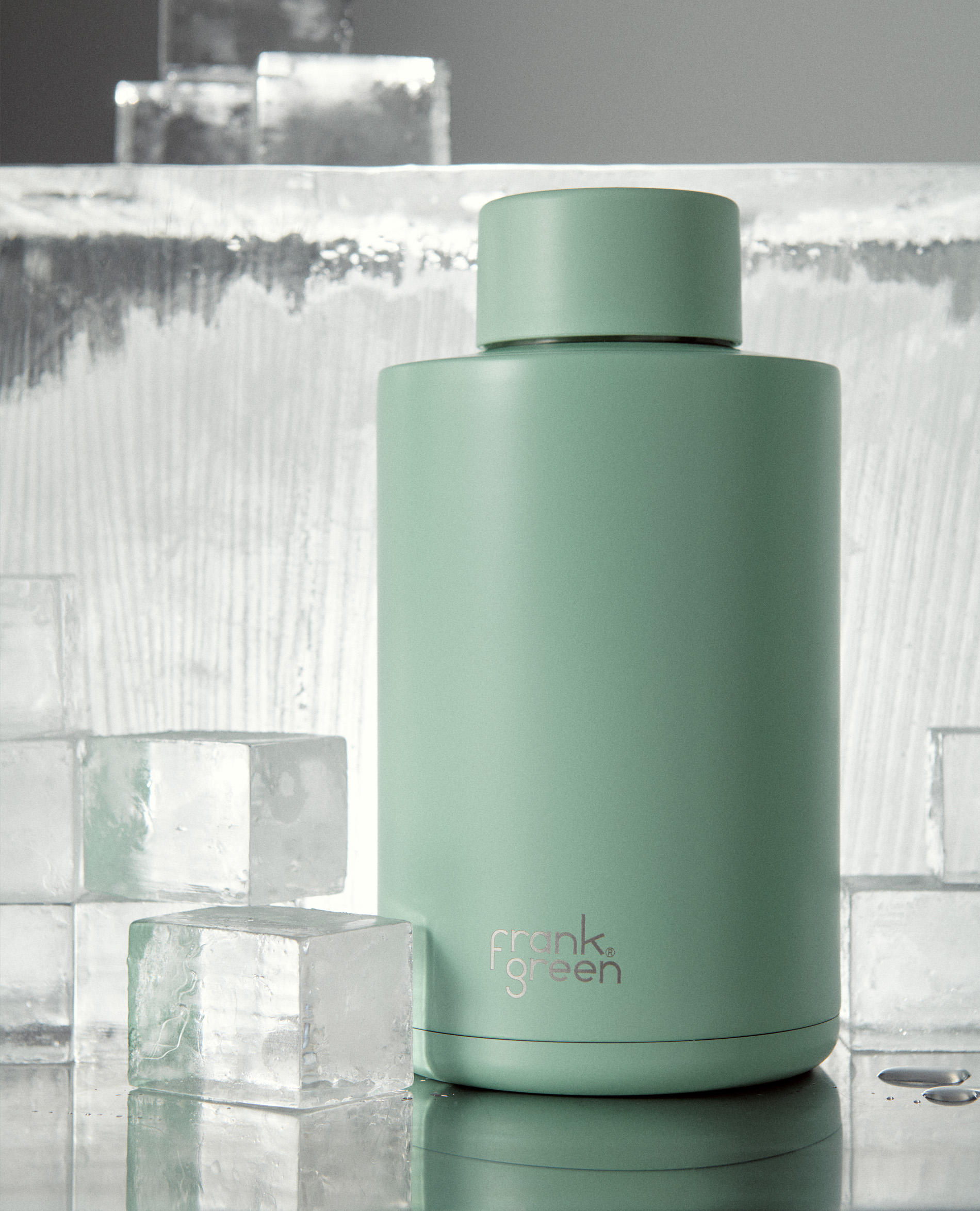 Mint green Frank Green water bottle on a reflective surface, surrounded by ice cubes and a wall of carved ice. Product photography.