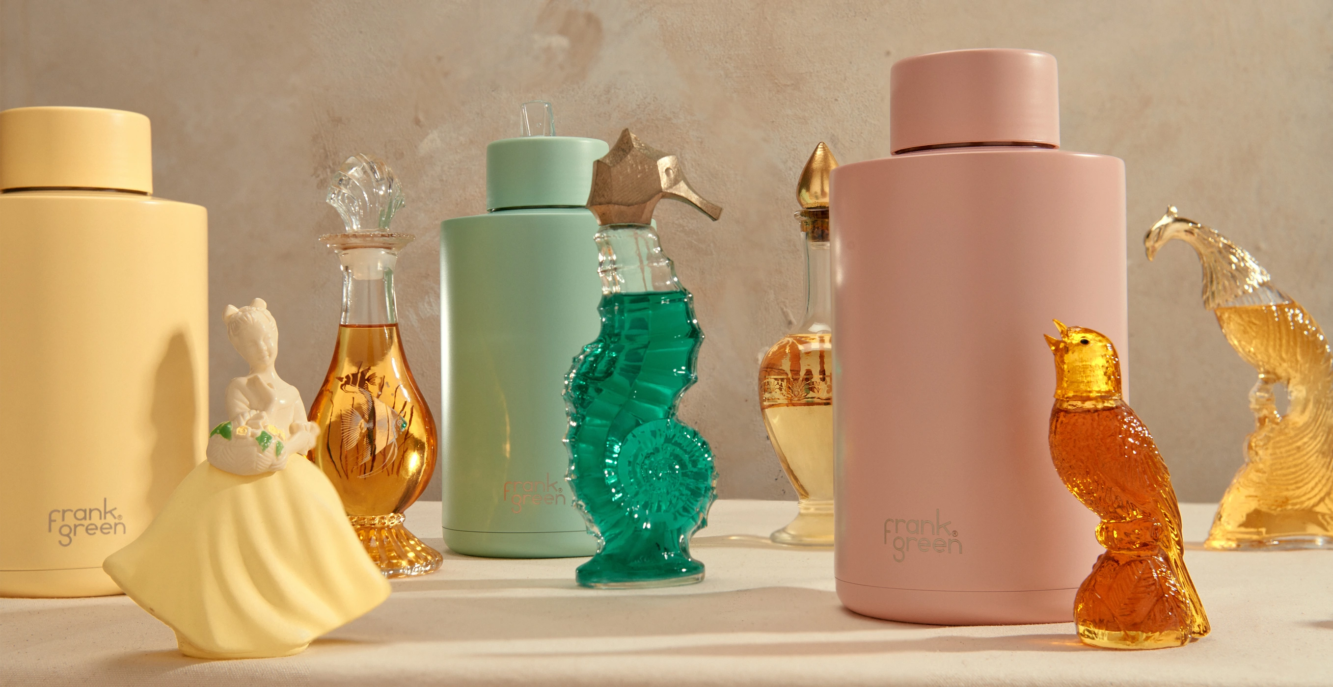 Frank Green reusable water bottles in pastel pink, mint, and yellow styled with vintage glass and ceramic figurines on a tabletop.