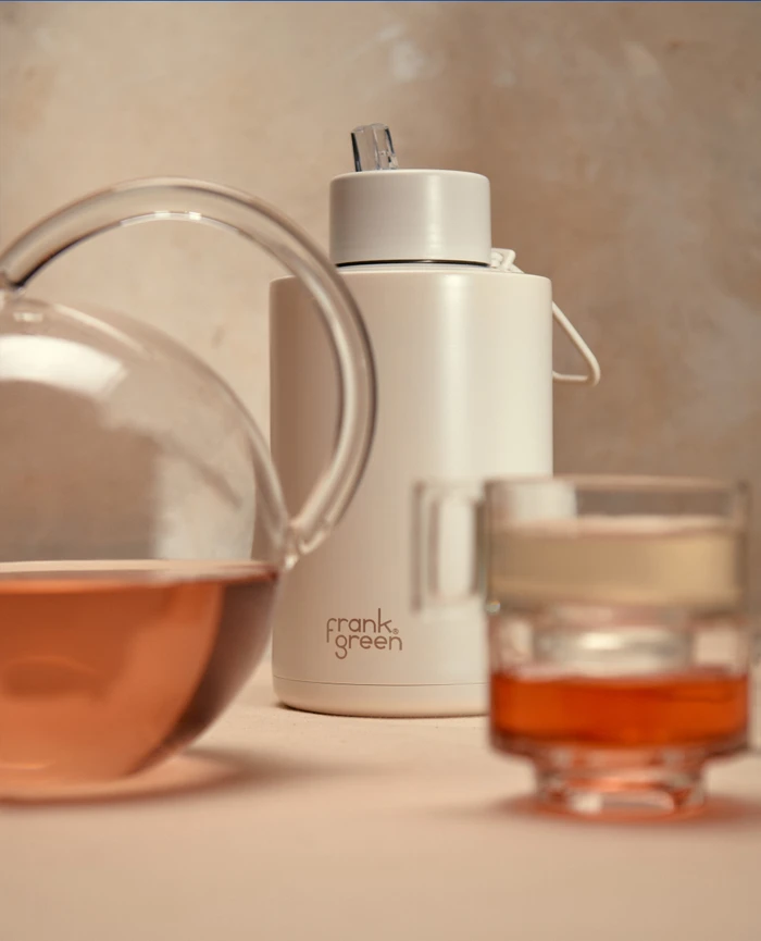 Off-white Frank Green reusable water bottle positioned between a clear glass teapot and teacup on a warm neutral surface.