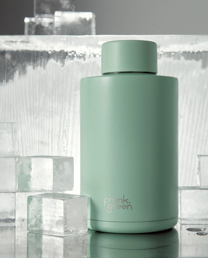 A mint green Frank Green reusable bottle standing upright surrounded by clear ice cubes on a reflective surface.