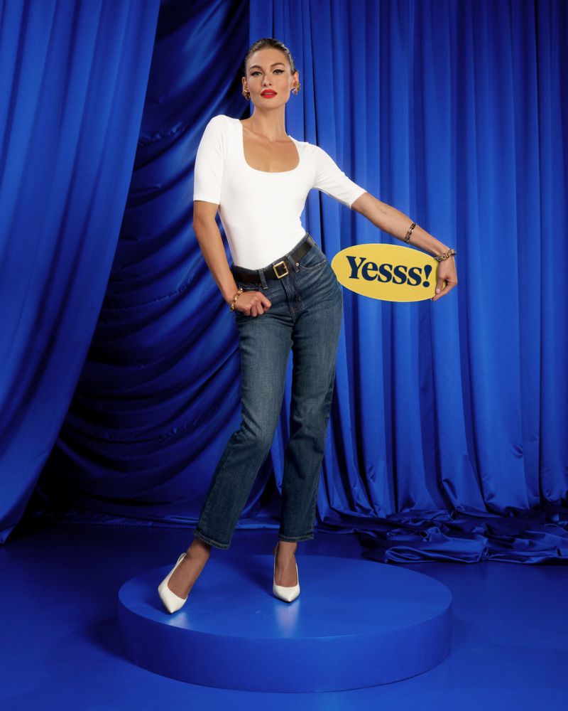 A woman posing on a blue platform in jeans, a white top, and white heels holds a yellow sign in a fashion photography studio.
