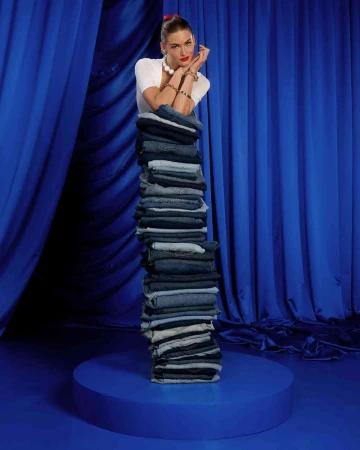 Young woman in white top and jewelry poses behind a tall stack of denim jeans on a blue circular platform with matching background drapes.