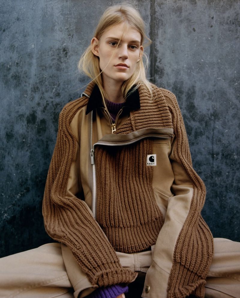 A blonde woman in a Carhartt jacket poses against a dark wall, showcasing a blend of knit and canvas textures with casual style.