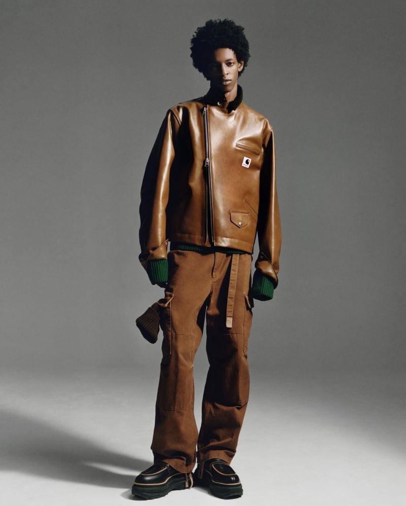 An adult Black male poses in a studio wearing a brown leather jacket, brown pants, and black shoes, illuminated by studio lighting.