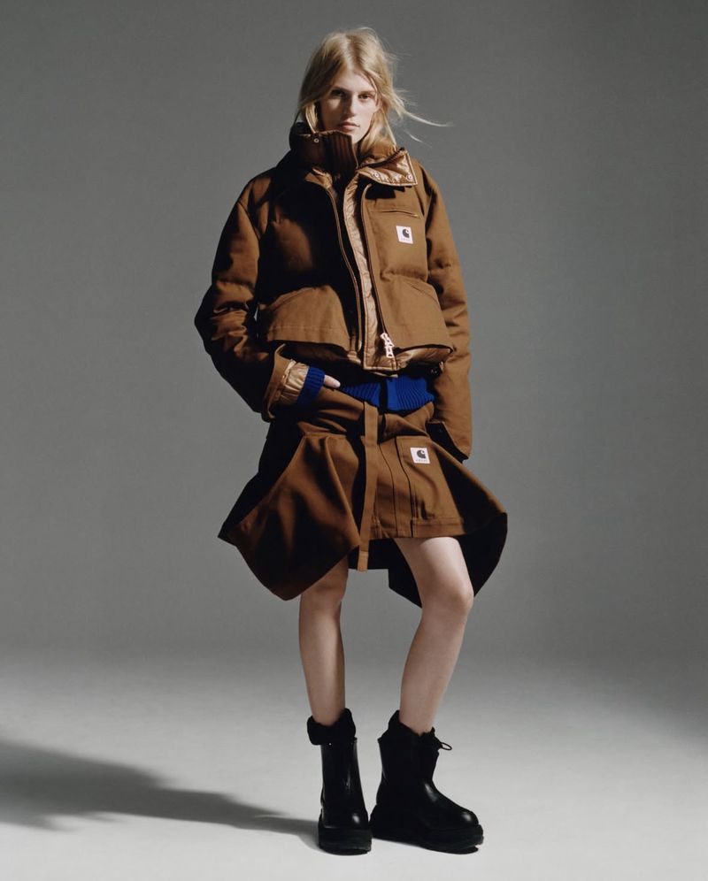 Fashion shot of a blonde woman in a brown Carhartt puffer jacket and skirt, paired with black boots, posing against a grey backdrop.