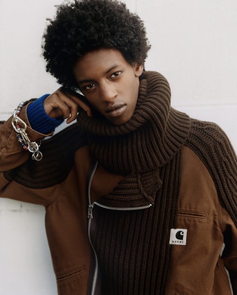 Man in a brown knit scarf and jacket poses indoors. The jacket has a Carhartt patch. He wears a blue sweater under the jacket.