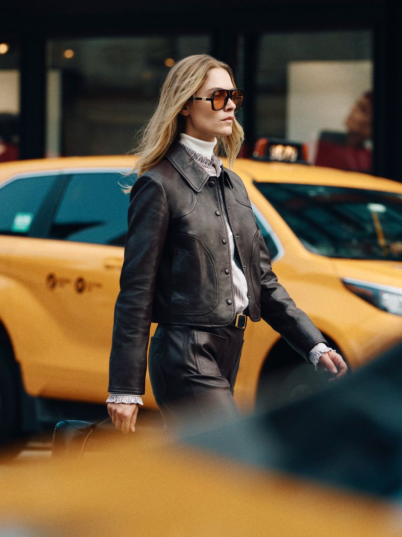 Stylish woman in a brown leather jacket walking on a city street with yellow taxi cabs, wearing sunglasses and a turtleneck.