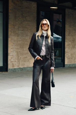 A full shot of a fashionable blonde woman in brown leather jacket and pants standing in front of a brick building, holding a black purse.