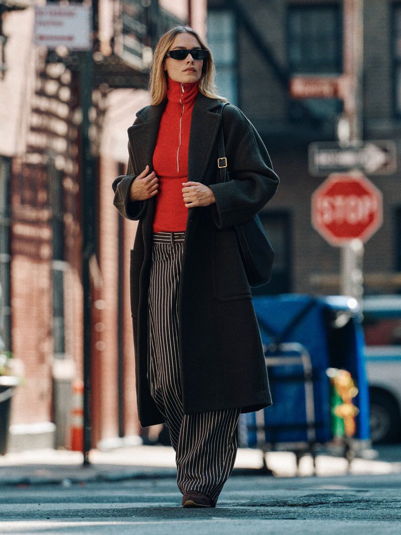Woman in coat walking on urban street with striped pants, red turtleneck, and sunglasses, with buildings and a stop sign in the background.