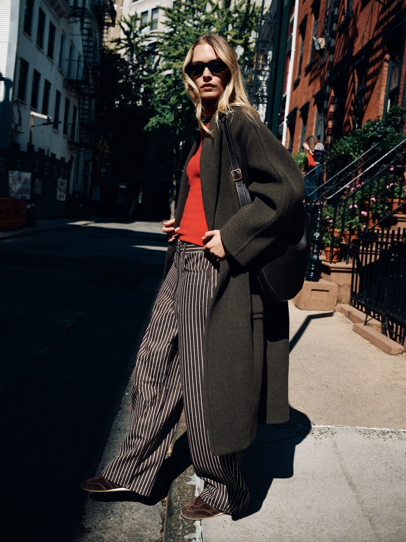 Model wearing a long dark olive coat, red knit top, and pinstriped trousers with sunglasses on a sunlit city street corner.