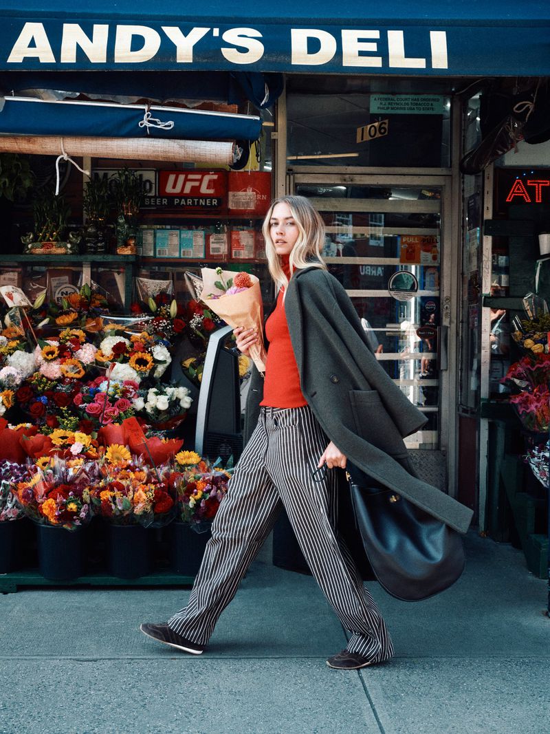 Adult woman walking in urban setting with flowers and a coat outside Andy's Deli, showcasing fashion and urban lifestyle.