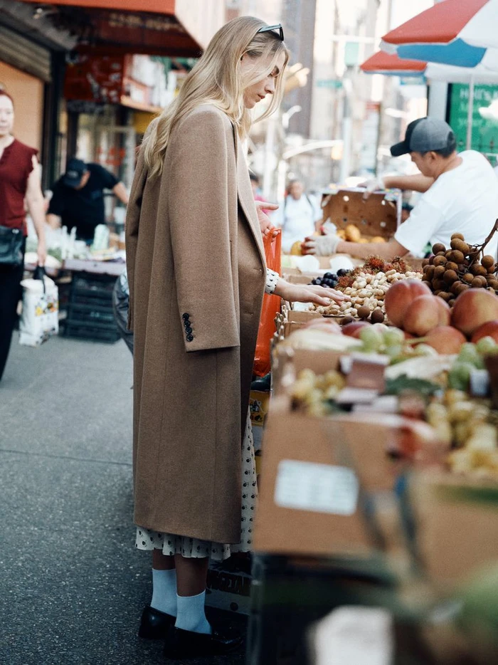 Blonde woman in a tailored camel coat and white socks examining produce at an outdoor fruit stand on a busy city sidewalk.