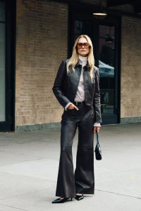 Model wearing a black leather jacket, wide-leg leather trousers, and oversized tinted sunglasses standing outside a brick building.