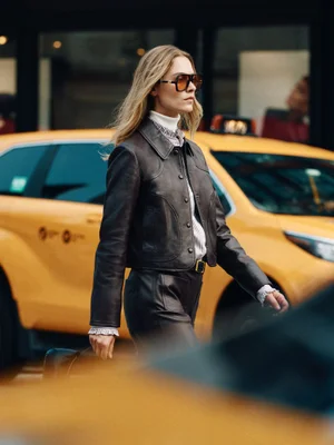 Model wearing a dark brown leather jacket, leather trousers, and oversized sunglasses walking past a yellow taxi on a city street.