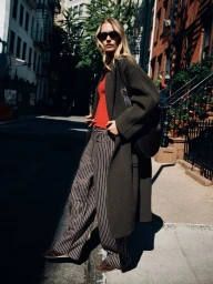 Model wearing a long dark olive coat, red knit top, and pinstriped trousers with sunglasses on a sunlit city street corner.