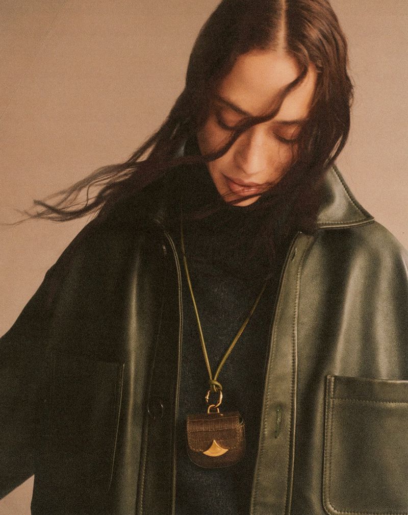 Woman in green leather jacket with brown purse necklace in studio. Dark hair. Fashion shot. Soft lighting. Neutral background.