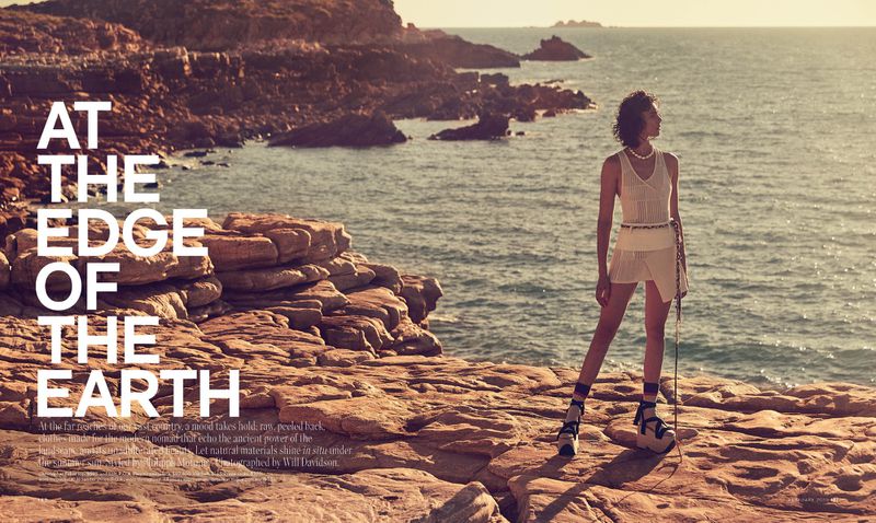 Woman stands on a rocky cliff edge overlooking the ocean in a fashion editorial shot at sunset with warm lighting and earthy tones.