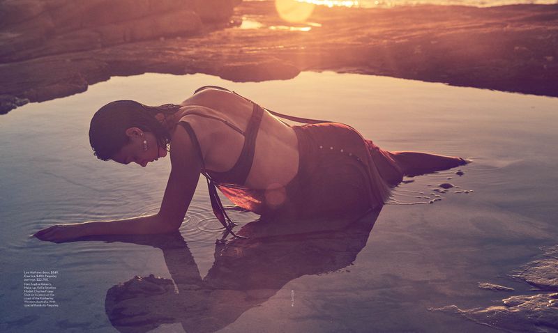 Woman in red dress laying in shallow pool of water at sunset, with rocky coastline and sun flares in background, creating a serene scene.