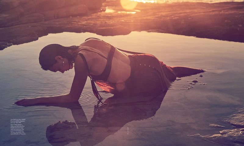 Woman in red dress laying in shallow pool of water at sunset, with rocky coastline and sun flares in background, creating a serene scene.