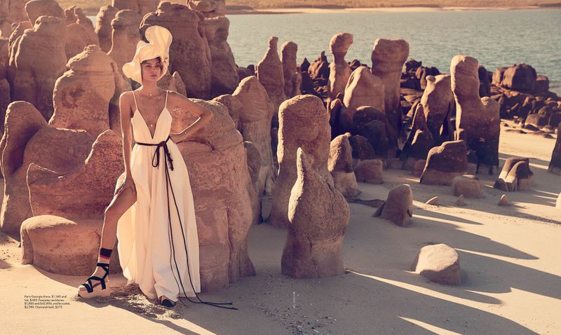 Model Charlee Fraser wearing a white dress and sculptural hat among sandy rock formations