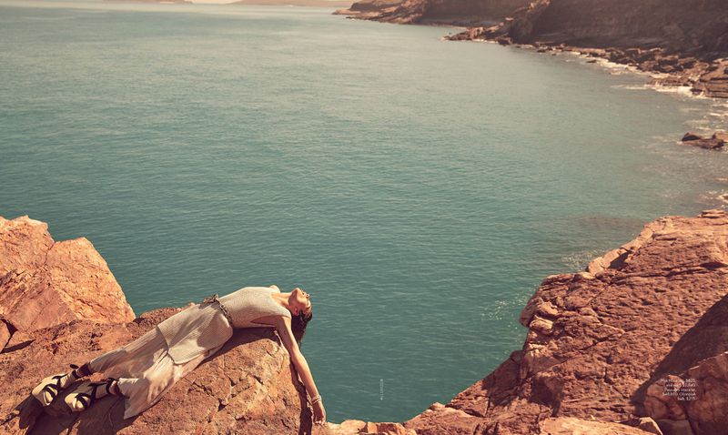 A woman in a dress lies on a rocky cliff overlooking turquoise ocean water with the rocky coastline in the background during daytime.