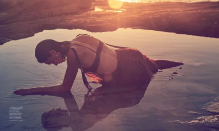 Model lying in shallow coastal tidal pool at sunset with warm golden light reflecting off the water surface