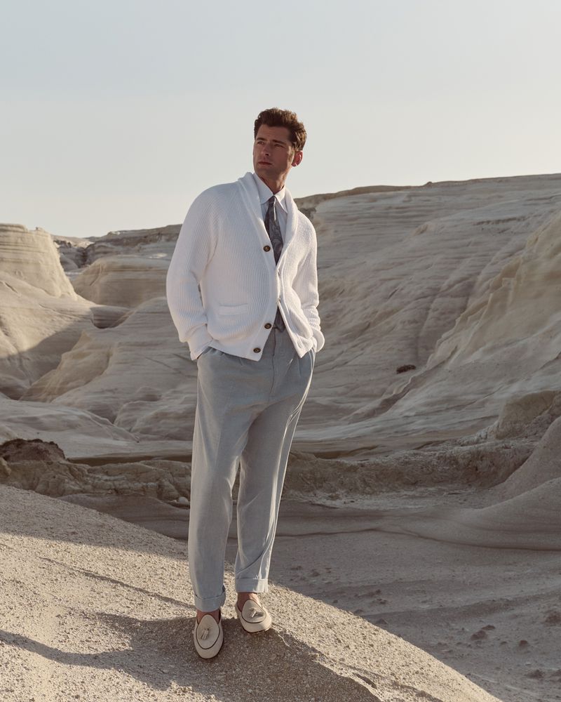 Model Sean O'Pry in a white knit cardigan and light trousers standing outdoors in a sandy desert landscape.