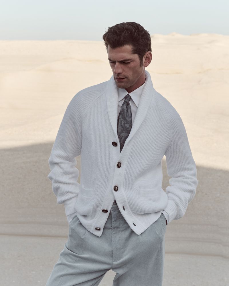Male fashion model poses in a white cardigan, dress shirt, and trousers outdoors in a desert-like setting with a neutral color palette.