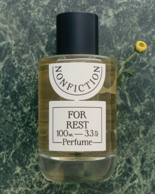 A bottle of Nonfiction perfume labeled 'For Rest' is displayed on a green marble surface, accompanied by a small yellow flower.