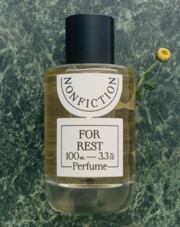 A bottle of Nonfiction perfume labeled 'For Rest' is displayed on a green marble surface, accompanied by a small yellow flower.