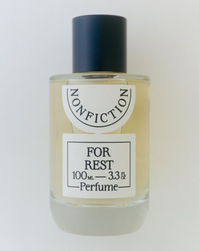 Close-up studio shot of Nonfiction For Rest perfume bottle with white label and blue cap on a neutral background, showcasing its design.