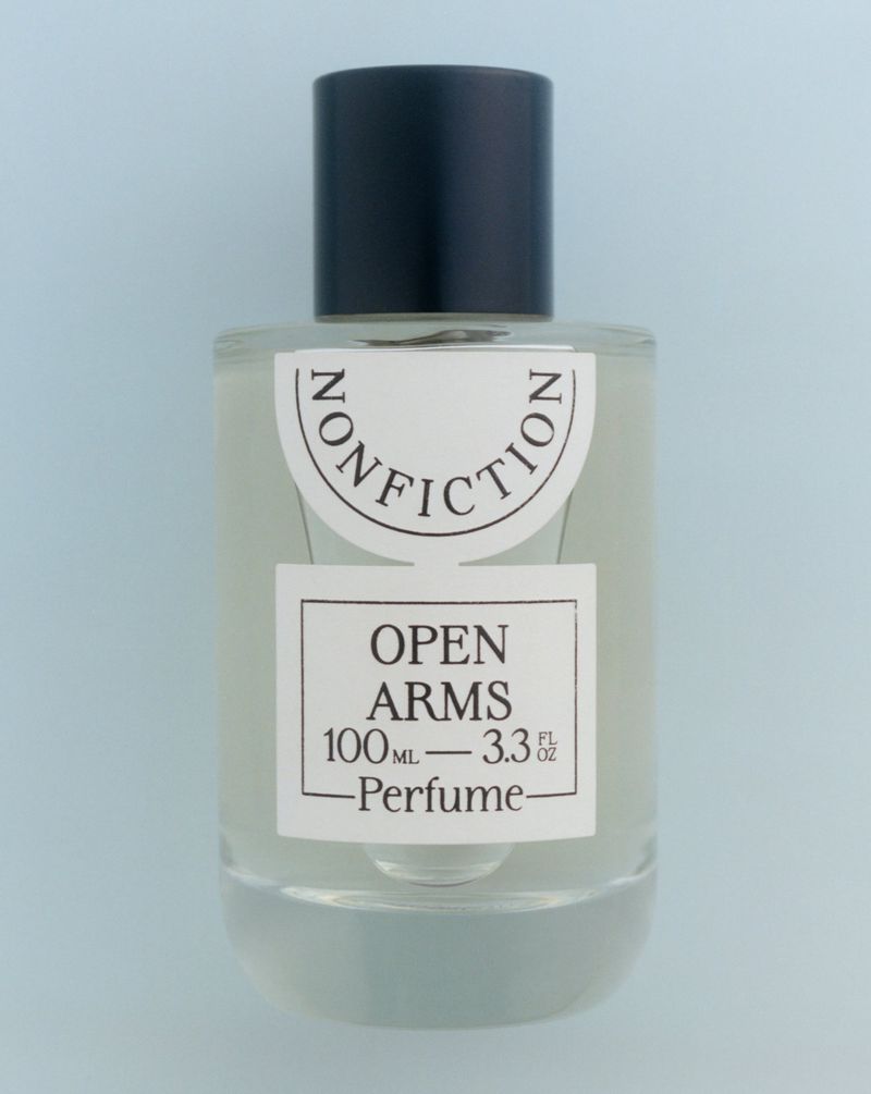 Clear glass bottle of Nonfiction Open Arms perfume with a black cap on a light blue background, centered and minimalist.