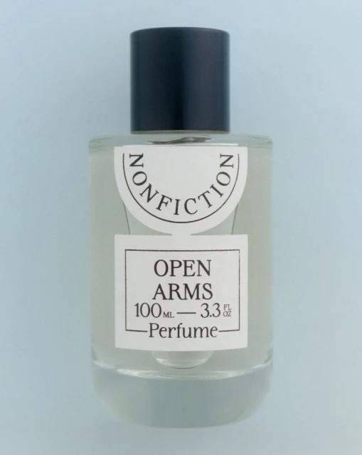 Clear glass bottle of Nonfiction Open Arms perfume with a black cap on a light blue background, centered and minimalist.