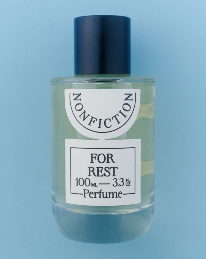 Studio shot of a Nonfiction For Rest perfume bottle with a black cap on a light blue background, showcasing its design and branding.