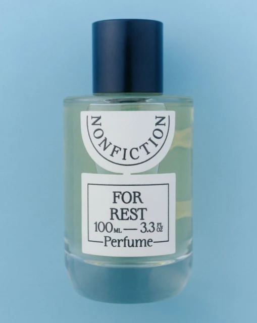 Studio shot of a Nonfiction For Rest perfume bottle with a black cap on a light blue background, showcasing its design and branding.