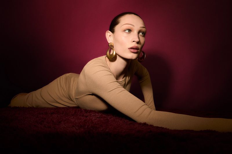 Fashion portrait of a young adult female lying on a red surface, wearing a beige dress and gold earrings against a maroon backdrop.