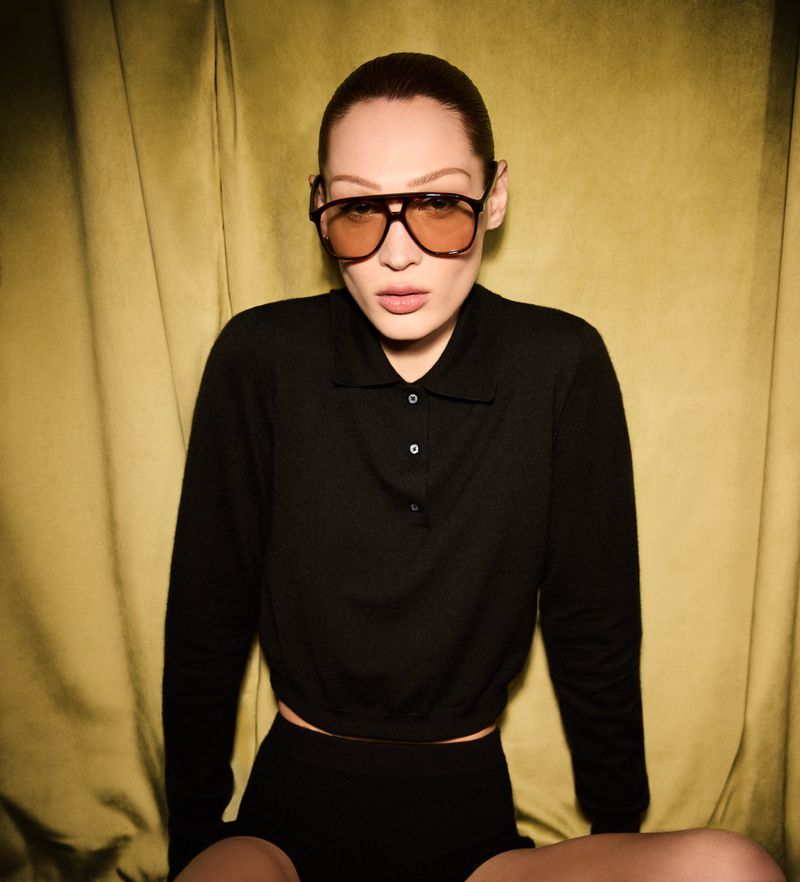 An adult woman in a dark cropped long-sleeved shirt and black shorts poses in front of a gold curtain wearing tinted aviator sunglasses.