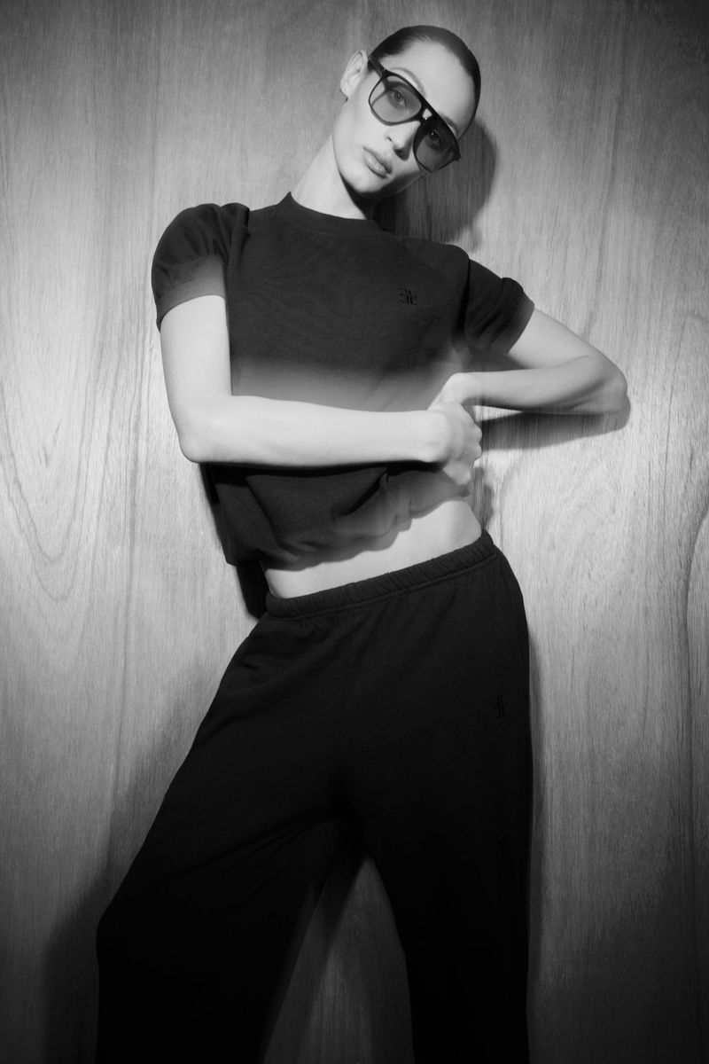 Monochrome portrait of a woman in a matching set and sunglasses posing against a wood panel background, in a studio setting.