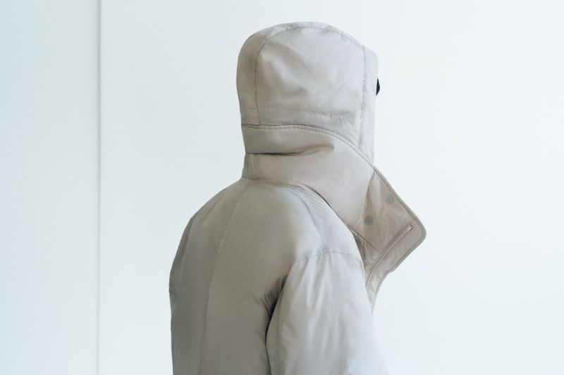 Back view of a person wearing a beige hooded down puffer jacket, standing against a plain white background in a neutral studio setting.