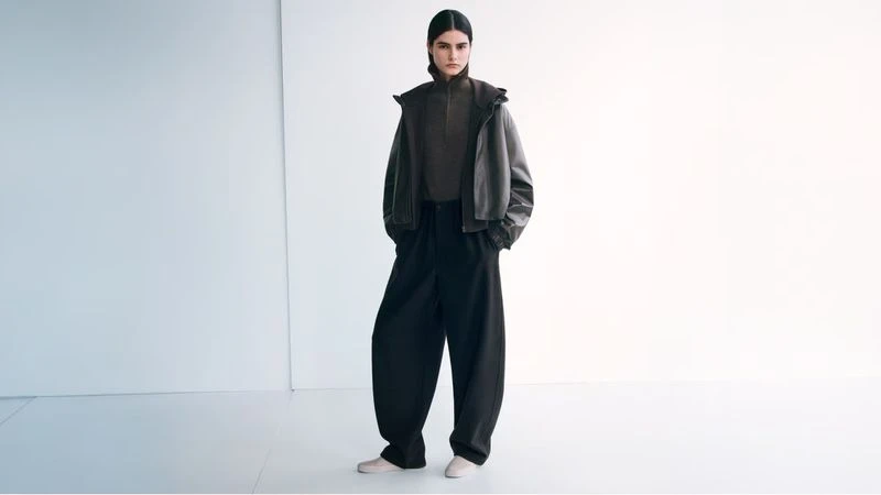 Fashion shot of a female model in black pants and a short leather jacket, posing in a bright white studio. Minimalist aesthetic.